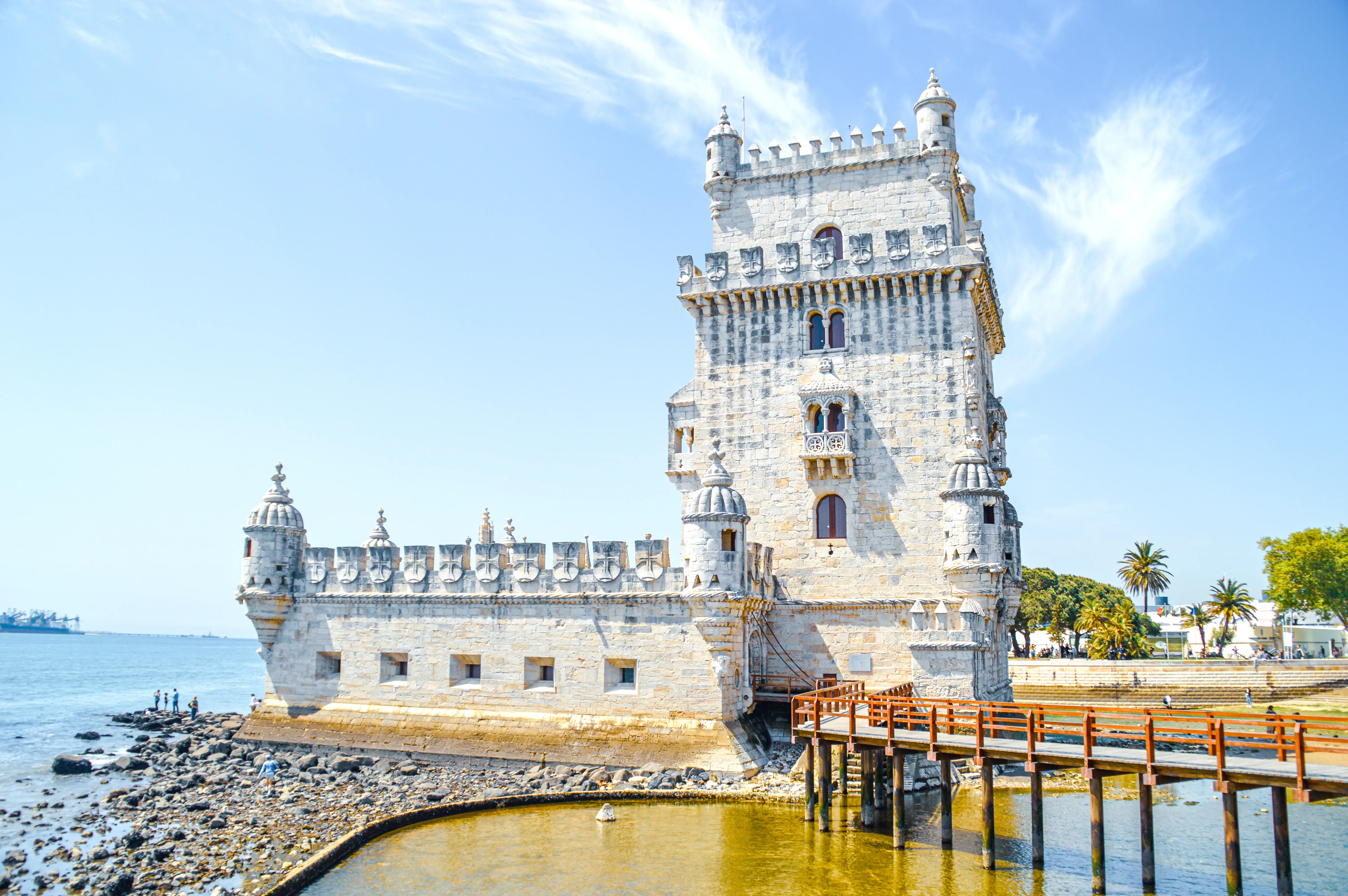 A wide-angle shot of the Belem Tower in Lisbon, Portugal, a stone fortress with ornate turrets and battlements, situated on the edge of the Tagus River. A wooden pier extends towards the tower from the right, and a rocky shoreline with a few people is visible on the left. The sky is clear blue with some thin clouds.